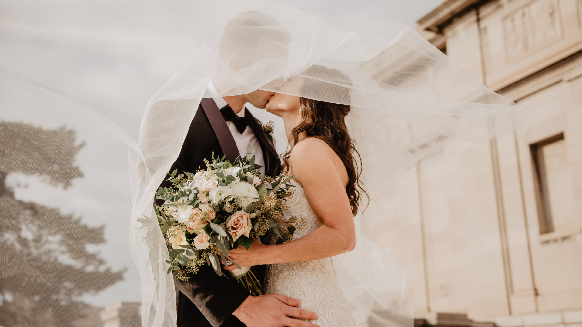 Novios besándose bajo un velo al viento, con un ramo de flores